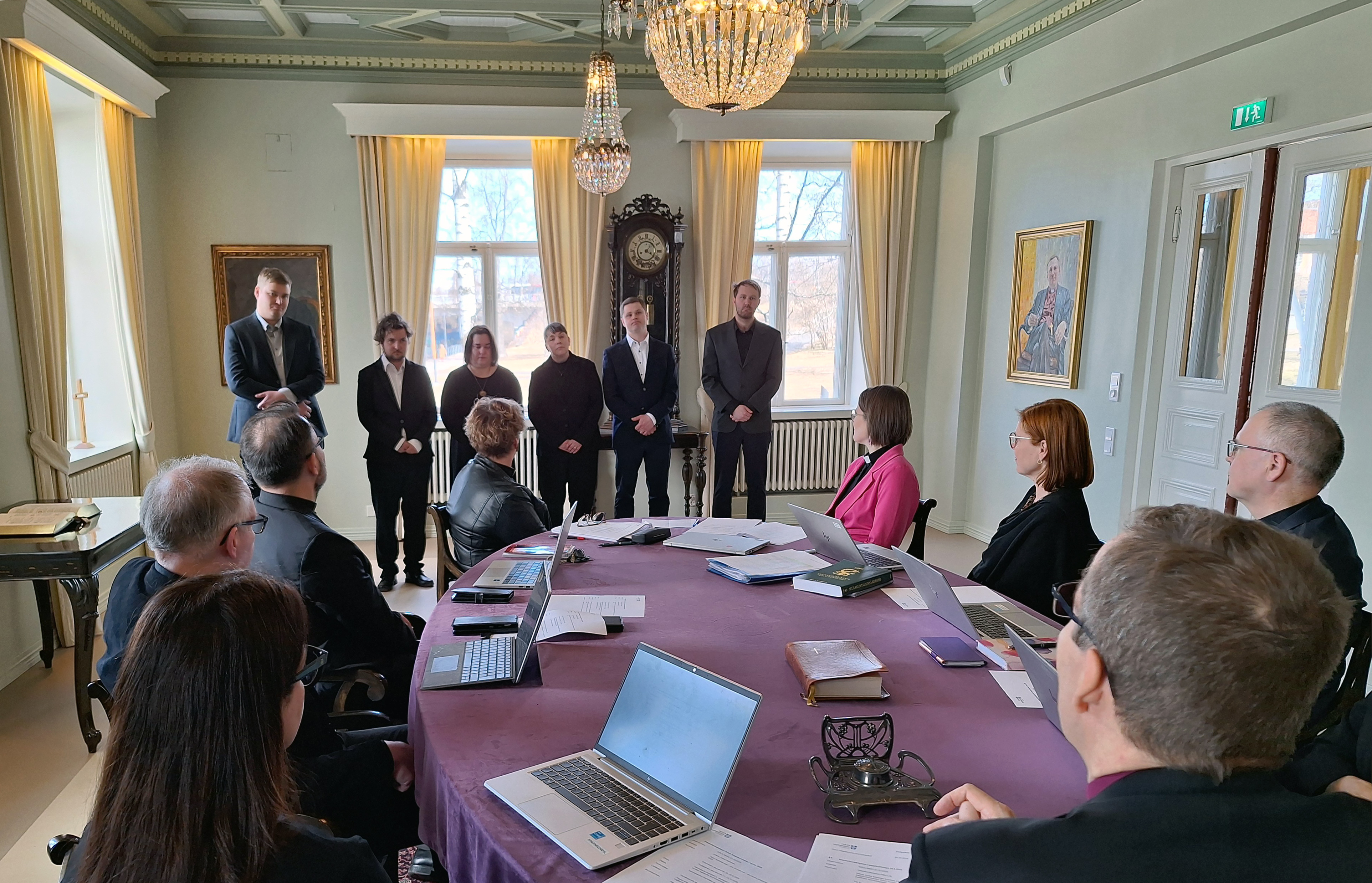 Ihmiset pöydän ympärillä katsovat kuutta rivissä seisovaa henkilöä, jotka saavat pian pappisvihkimyksen.
